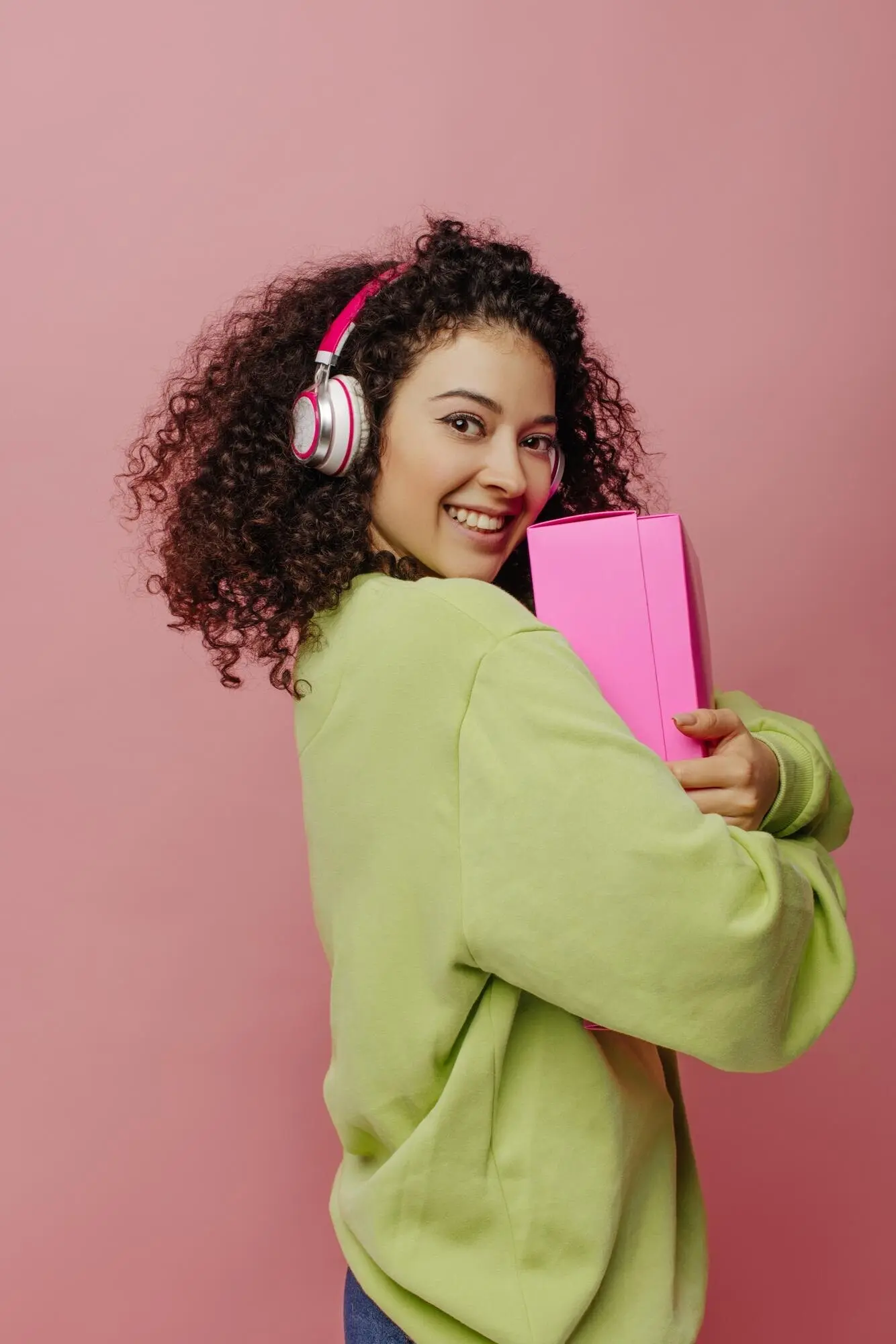 Eine attraktive, hellhäutige junge Frau steht seitlich und hält eine Box an sich vor pinkem Hintergrund; die Brünette mit Kopfhörern und Jacke lächelt in die Kamera. Konzept: Emotion, Gesichtsausdruck.